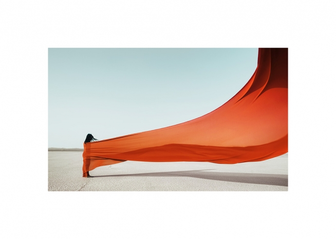  - Fotografi av en kvinne som er pakket inn i et oransje stoff som blafrer i vinden, mot en lyseblå bakgrunn
