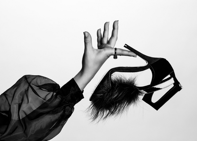  – Svarthvitt-fotografi av en høyhælt sko med pels på tåen, som holdes av en kvinne i en gjennomsiktig puffbluse