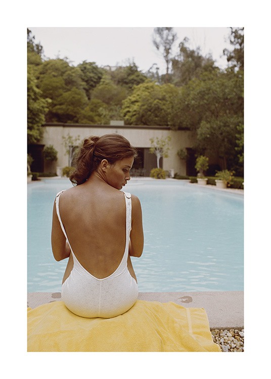  – Fotografi av en kvinne som sitter på et håndkle foran et basseng, iført en hvit badedrakt