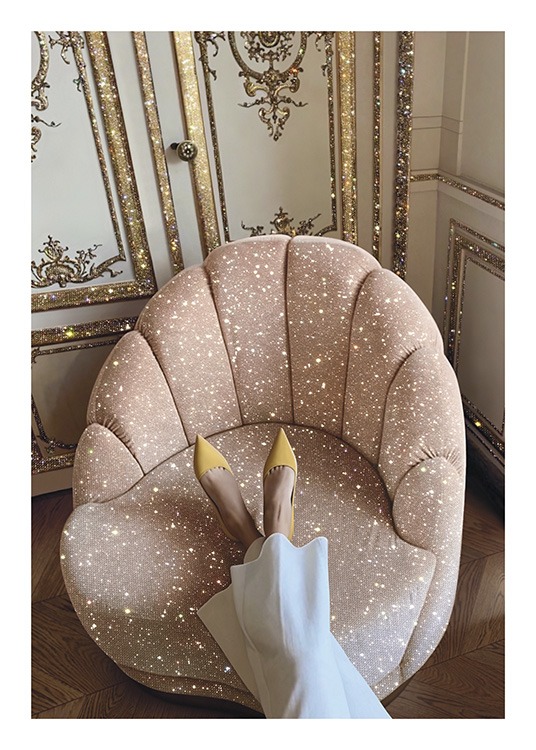  – Fotografi av en rosa, glitrende stol foran en gyllen og hvit vegg, med en kvinne som hviler foten på stolen